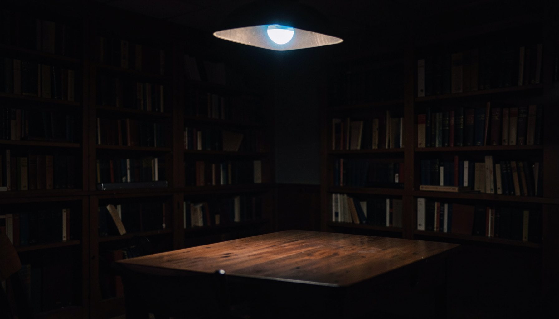 A dark, low-key photograph with deep brown and navy tones, showing a quiet corner of a library or study with soft overhead light