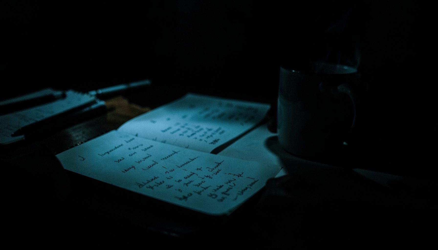 A dark, low-key photograph with rich charcoal and muted teal shadows, showing a desk with handwritten notes and a steaming mug