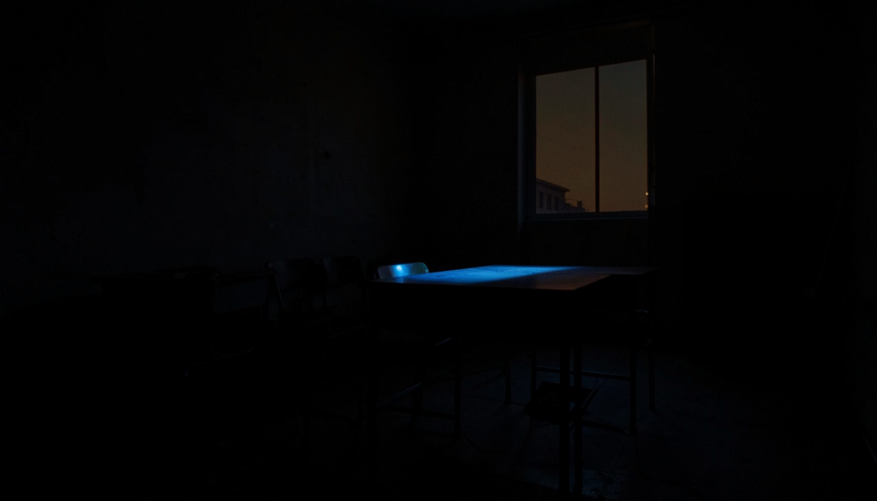 A dark, low-key photograph of an empty classroom at dusk, with deep shadows pooling in the corners and a single desk illuminated by soft window light