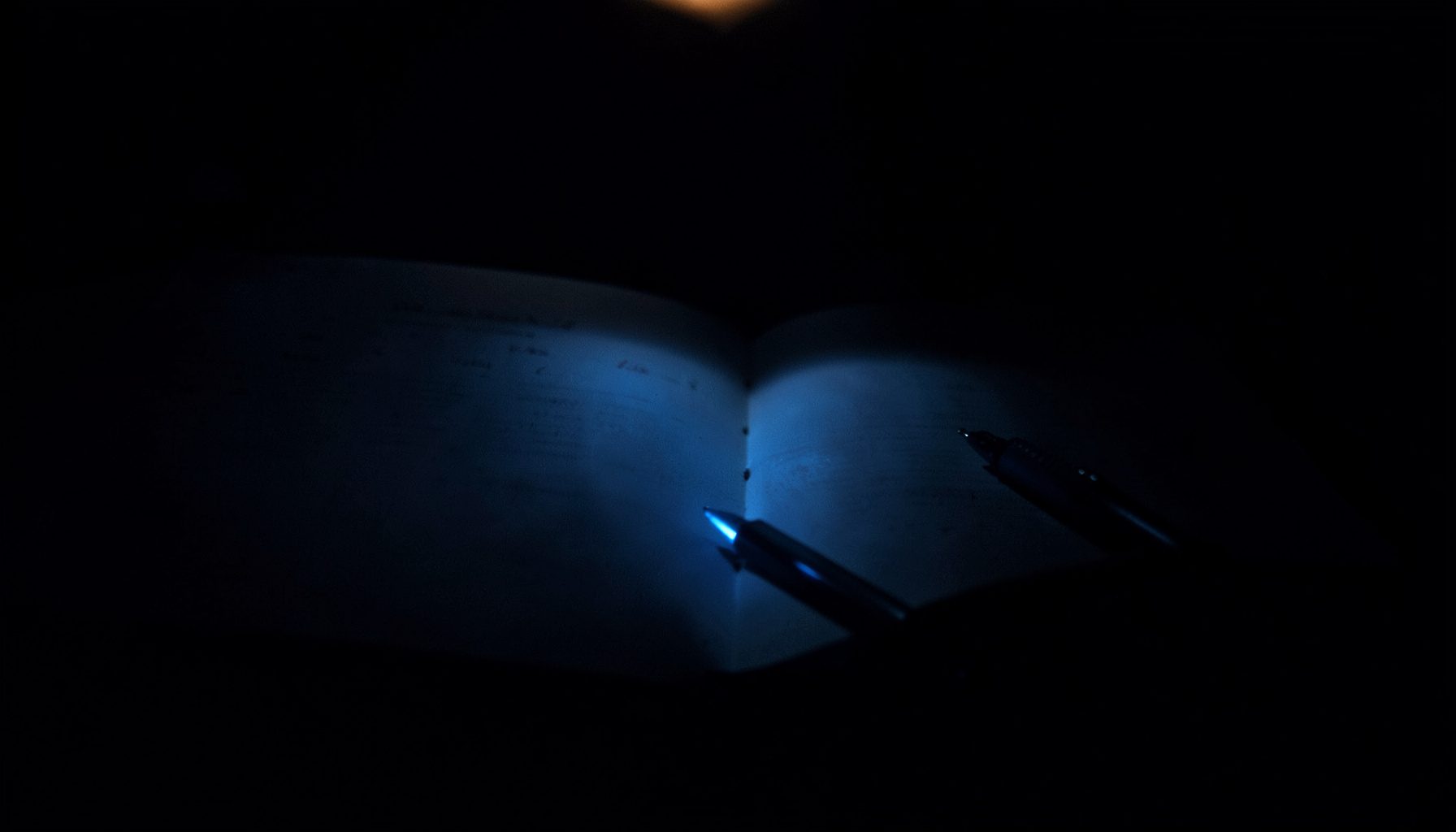 A dark, low-key photograph with deep indigo and charcoal shadows, showing an open journal beside a pen, lit by a single warm lamp