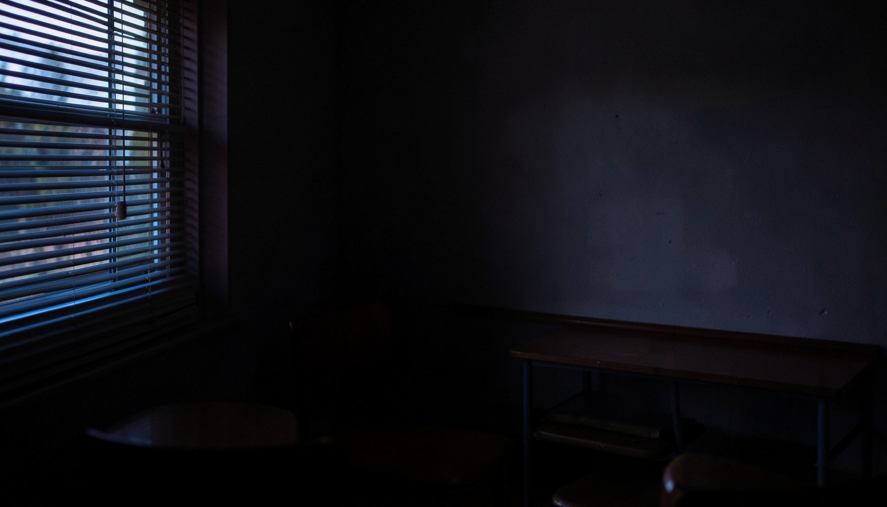 A dark, low-key photograph with rich charcoal and muted plum shadows, showing a quiet classroom corner with soft natural light