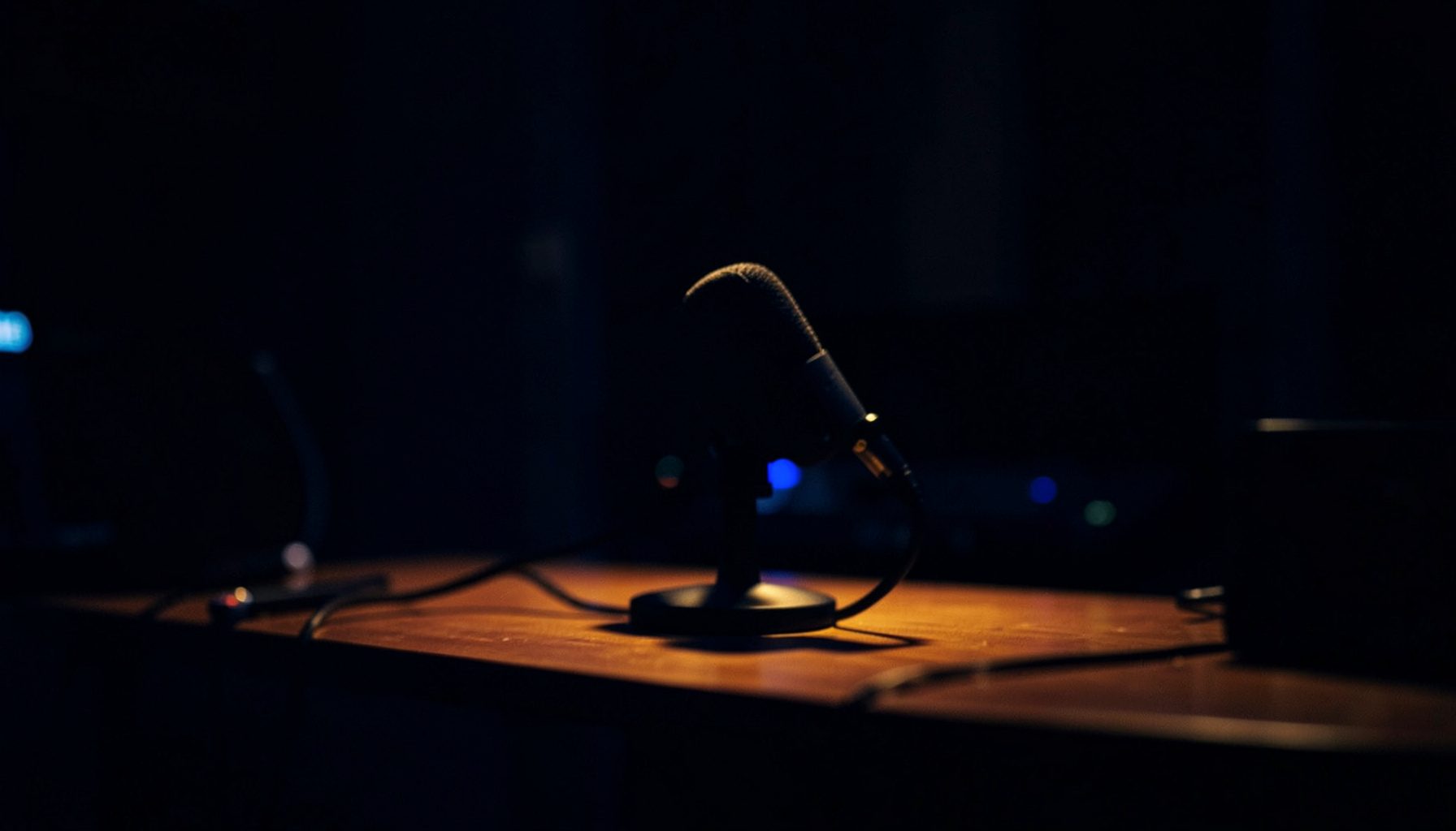 A dark, low-key photograph with deep navy and warm amber tones, showing a microphone on a desk in a softly lit recording studio