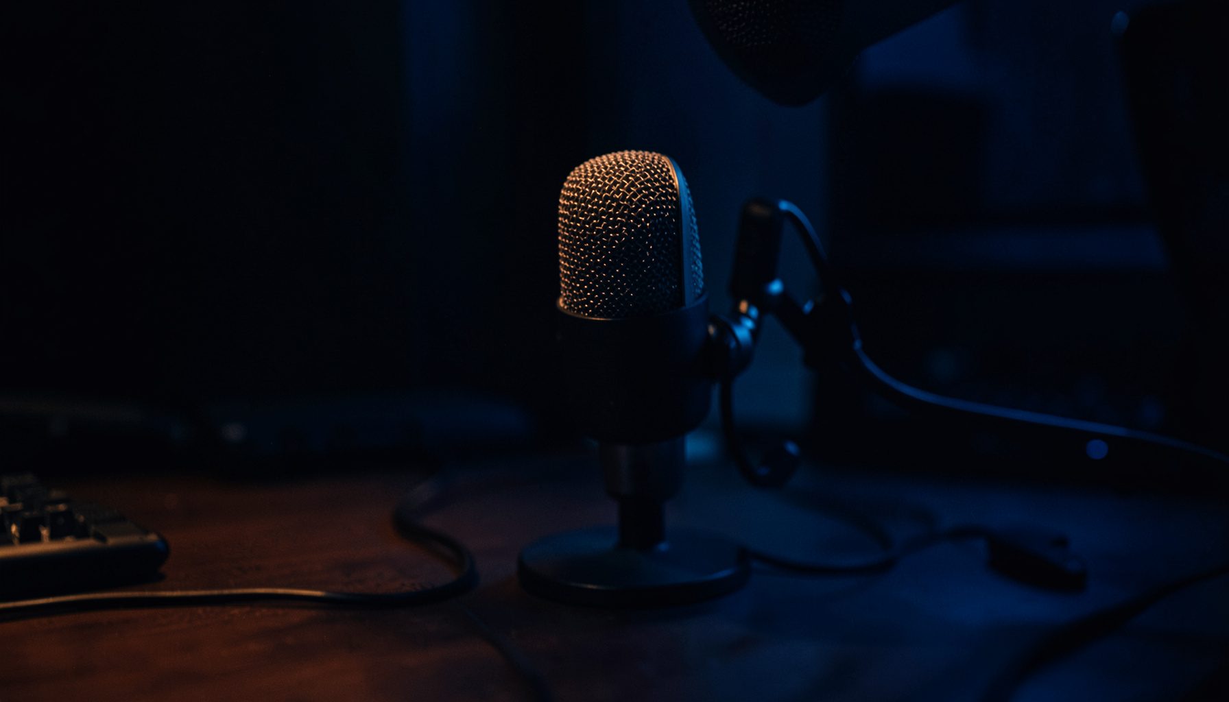 Dark, low-key photograph of a professional podcast microphone on a desk in a dimly lit studio, deep navy and charcoal tones