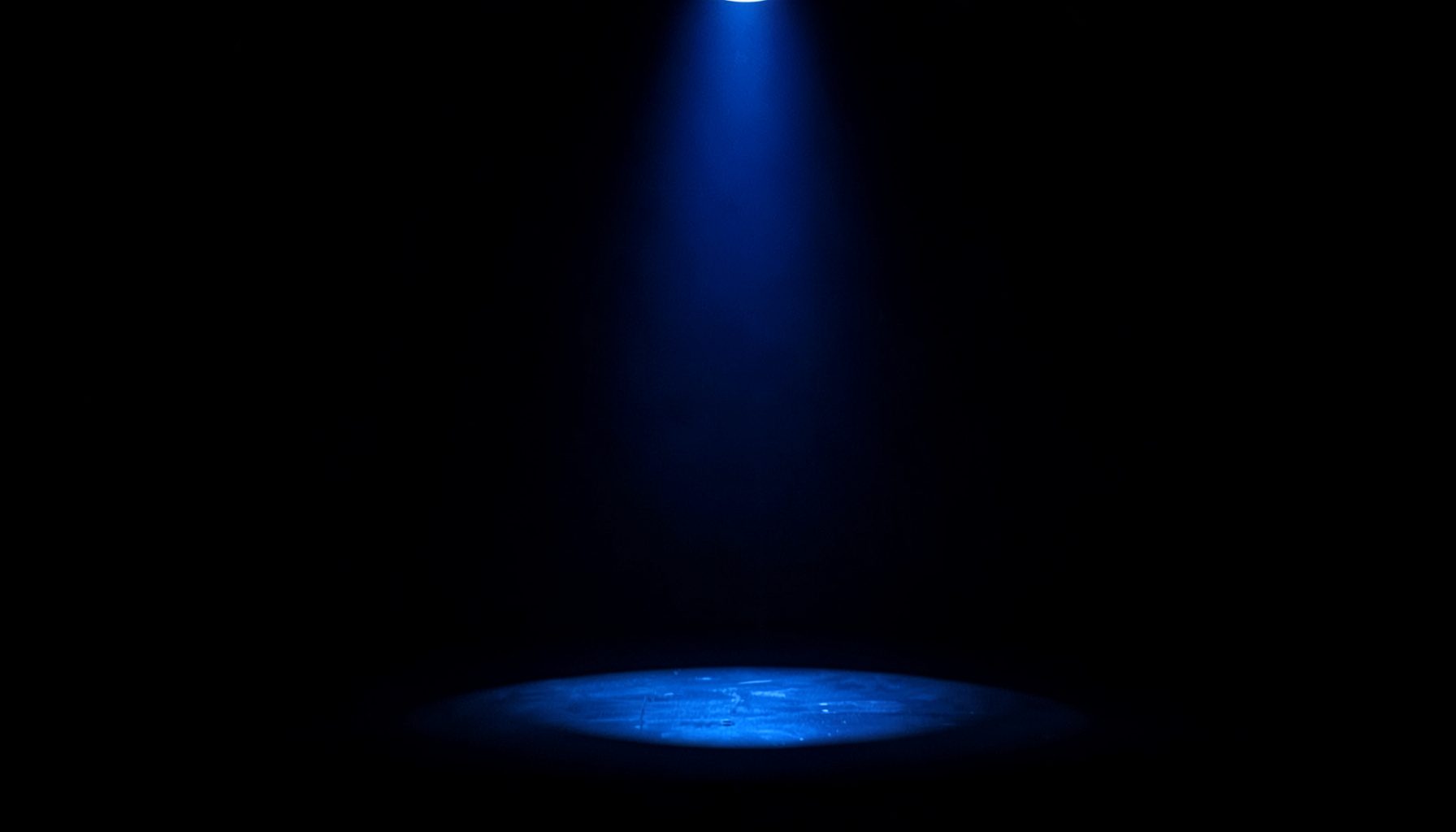 A dark, low-key photograph of a spotlight illuminating an empty stage with deep navy and charcoal shadows, evoking anticipation