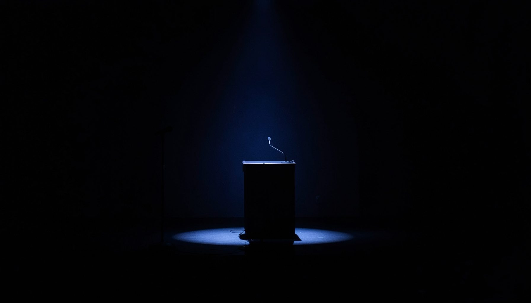 A dark, low-key photograph of a conference stage with a spotlight on an empty lectern, deep navy and charcoal shadows filling the room