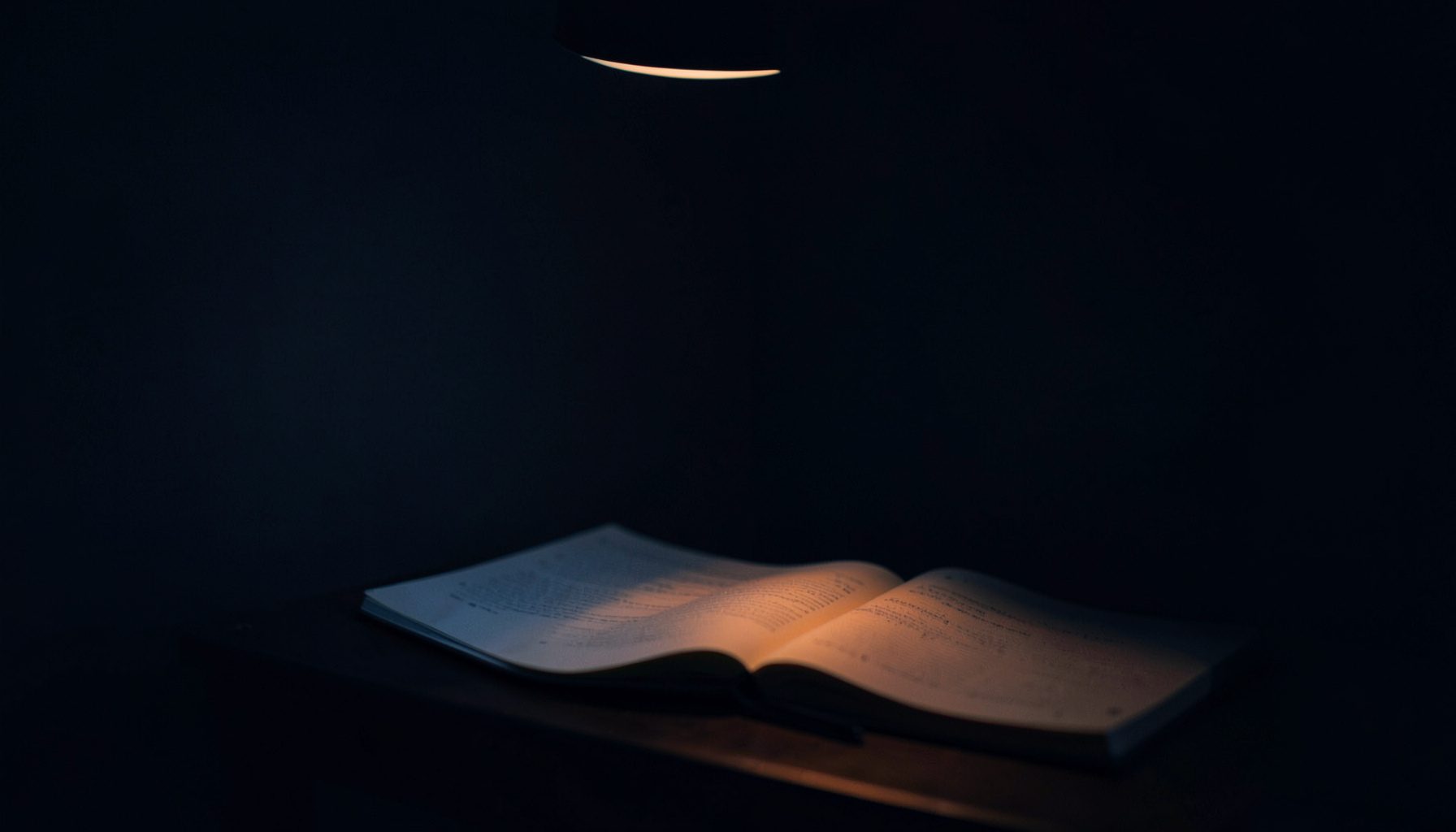 A dark, low-key photograph with deep navy and charcoal shadows, showing a quiet study corner with an open notebook and soft natural light