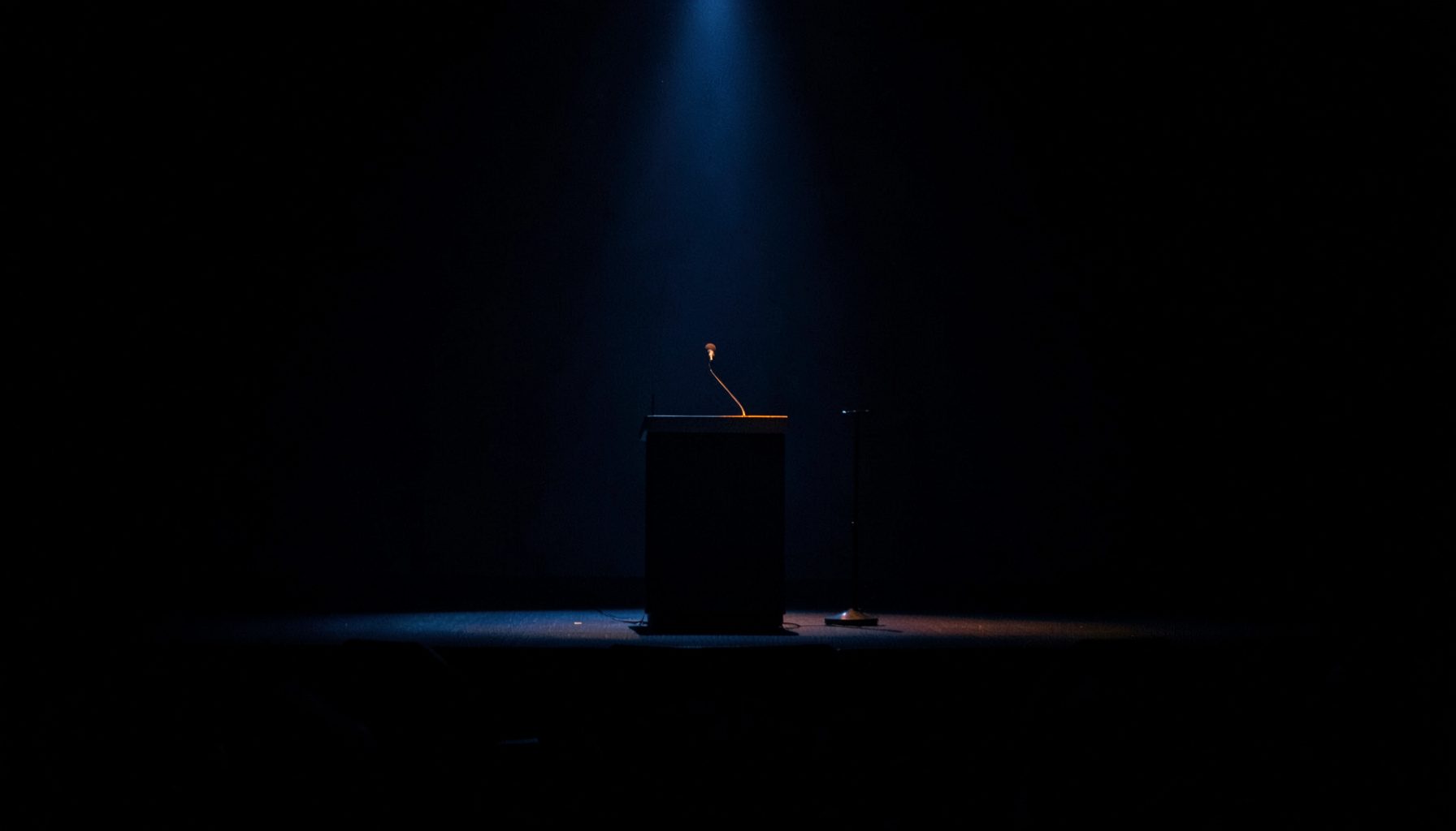 A dark, low-key photograph of a conference stage with a spotlight on an empty lectern, deep navy and charcoal shadows fill the frame