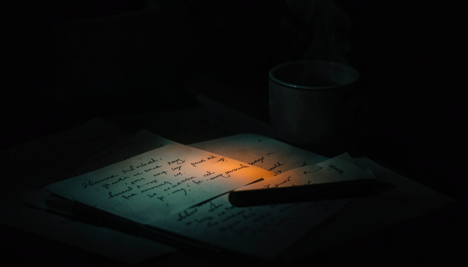 Dark, low-key photograph with rich charcoal and muted teal shadows, showing a desk with handwritten notes and a steaming mug