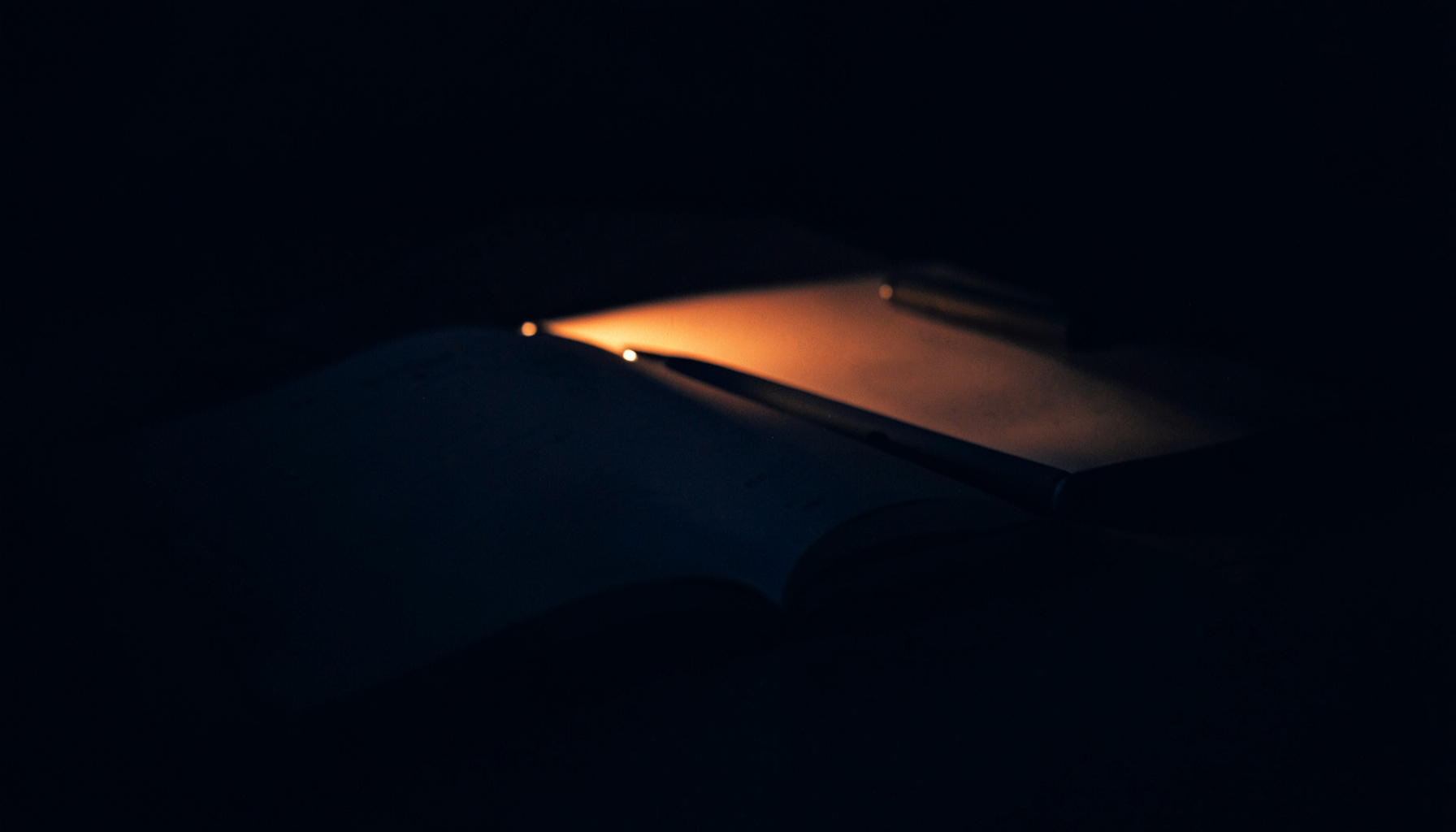 A dark, low-key photograph with deep indigo and charcoal shadows, showing an open journal beside a pen, lit by a single warm lamp